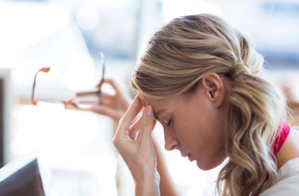 Young woman experiencing headaches at work while she takes off glasses to touch the pain