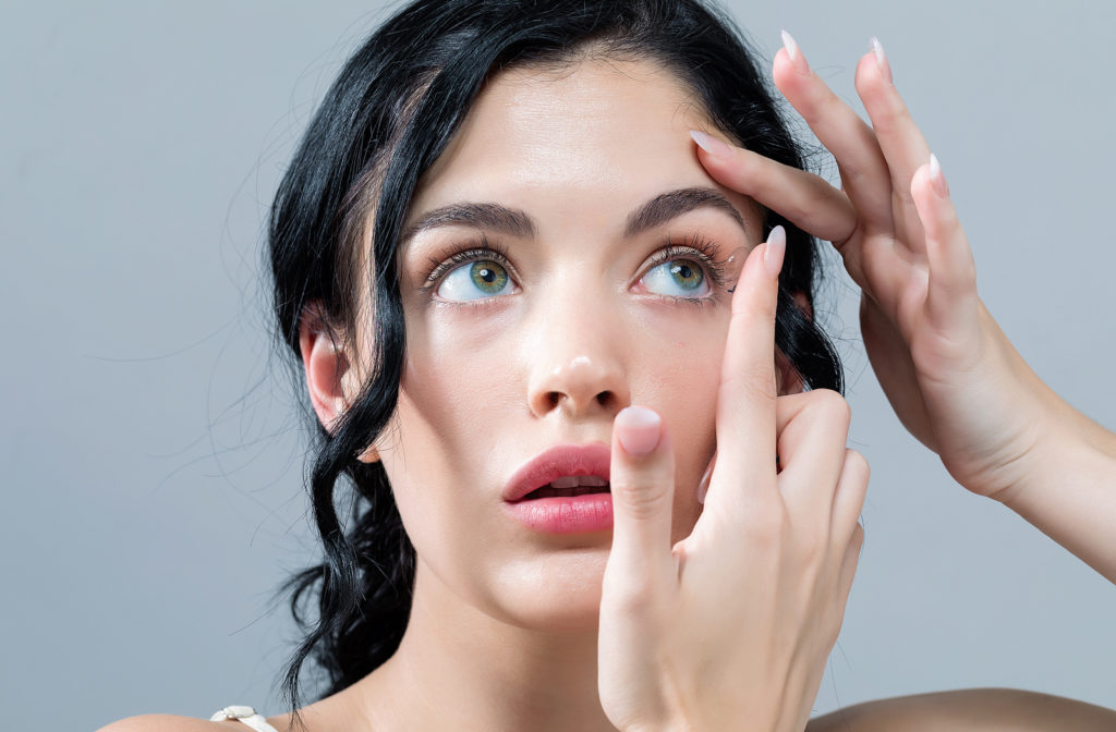 Young woman putting on contact lenses while she looks up on grey background