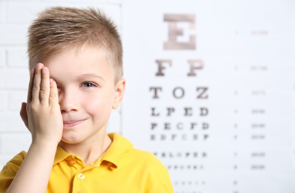 Young boy covering one eye to undergo eye exam with eye exam chart in background faded