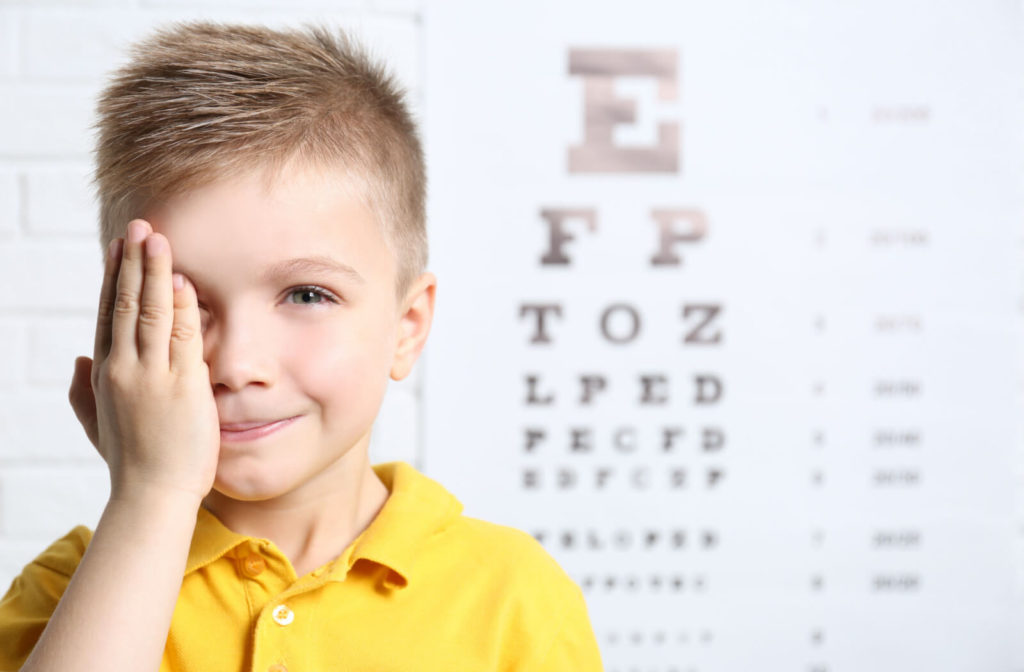 A boy in a yellow shirt is covering his right eye with his right palm and there's a Snellen chart in the background.