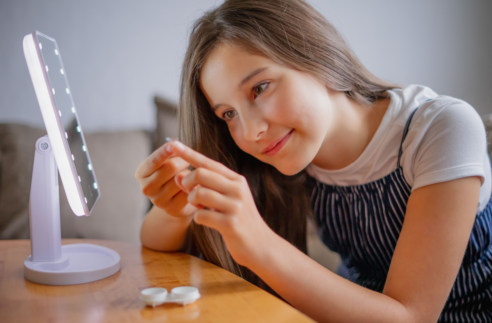 A young girl in front of a small mirror smiling while looking closely at an ortho-k lens between her fingers.
