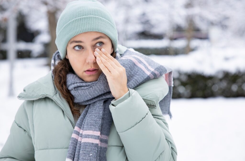 A woman, wrapped in winter clothing, touches beneath her eye outdoors, experiencing symptoms of dry eyes.