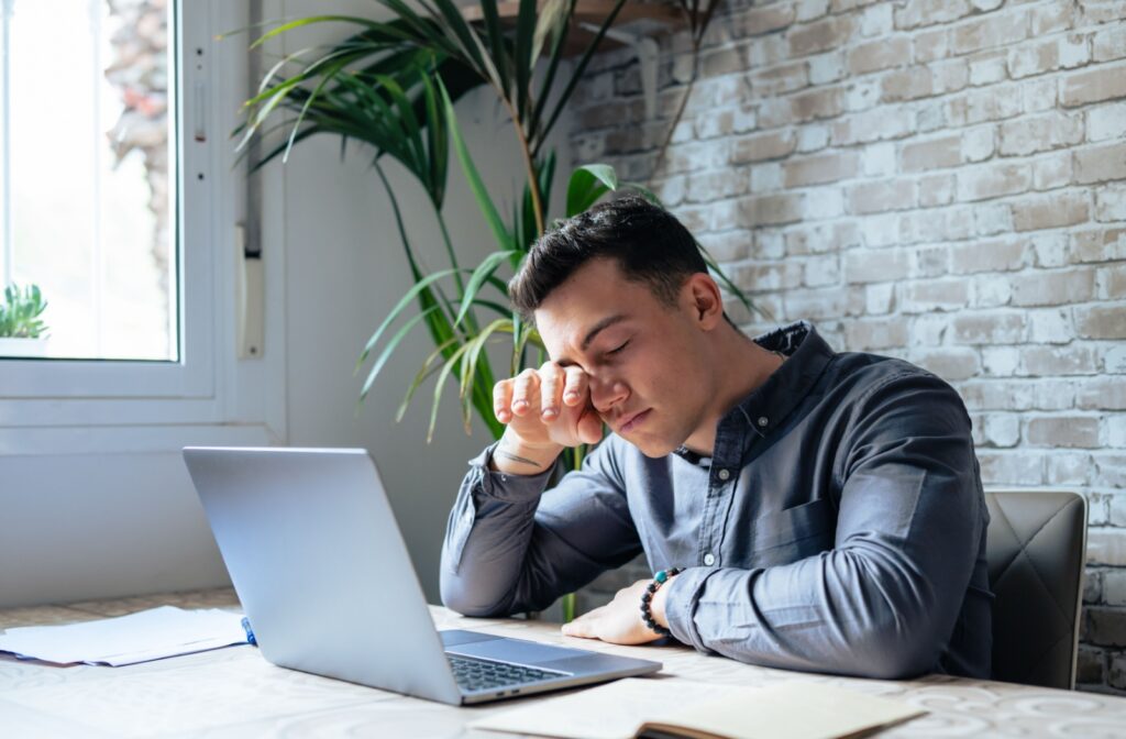 A man is sitting at a desk, rubbing his eyes in discomfort, likely due to eye strain or dryness.