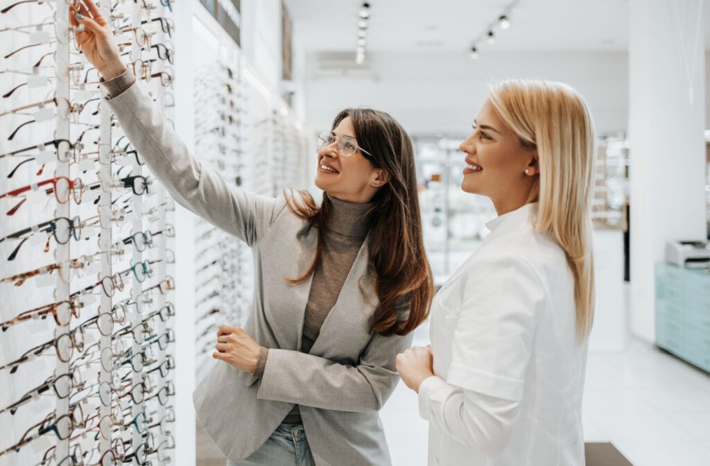 Two people stand by a wall of eyeglass frames, one reaching for a pair while the other smiles.