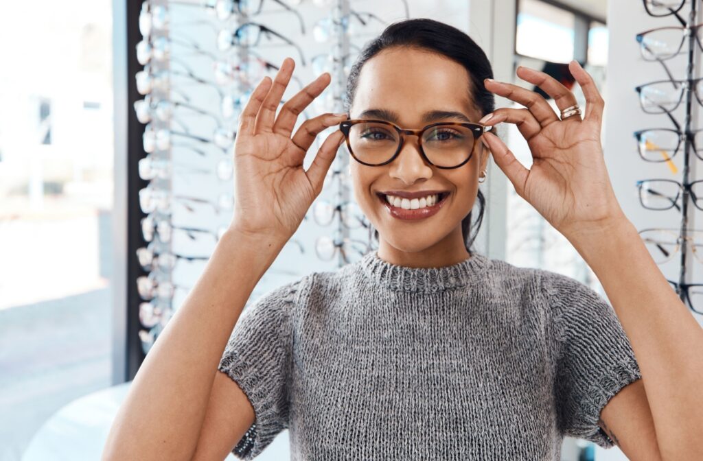 Smiling young adult wearing glasses in an optical store, holding onto glasses.