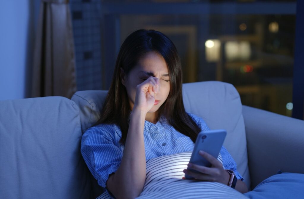 A person looking at their phone on the couch at night, rubbing their eye.